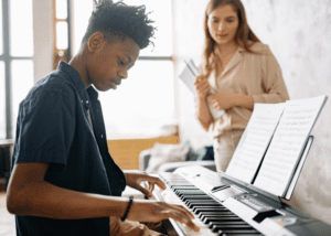 Boy practicing piano while female teacher is standing nearby