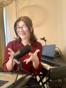 Woman sitting by digital piano talking into microphone on computer